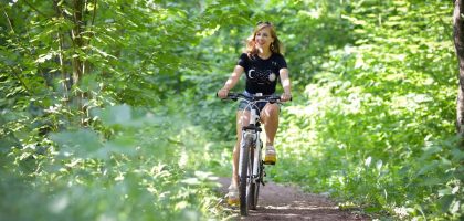 A woman cycling on a scenic forest trail surrounded by green foliage, enjoying a sunny day.